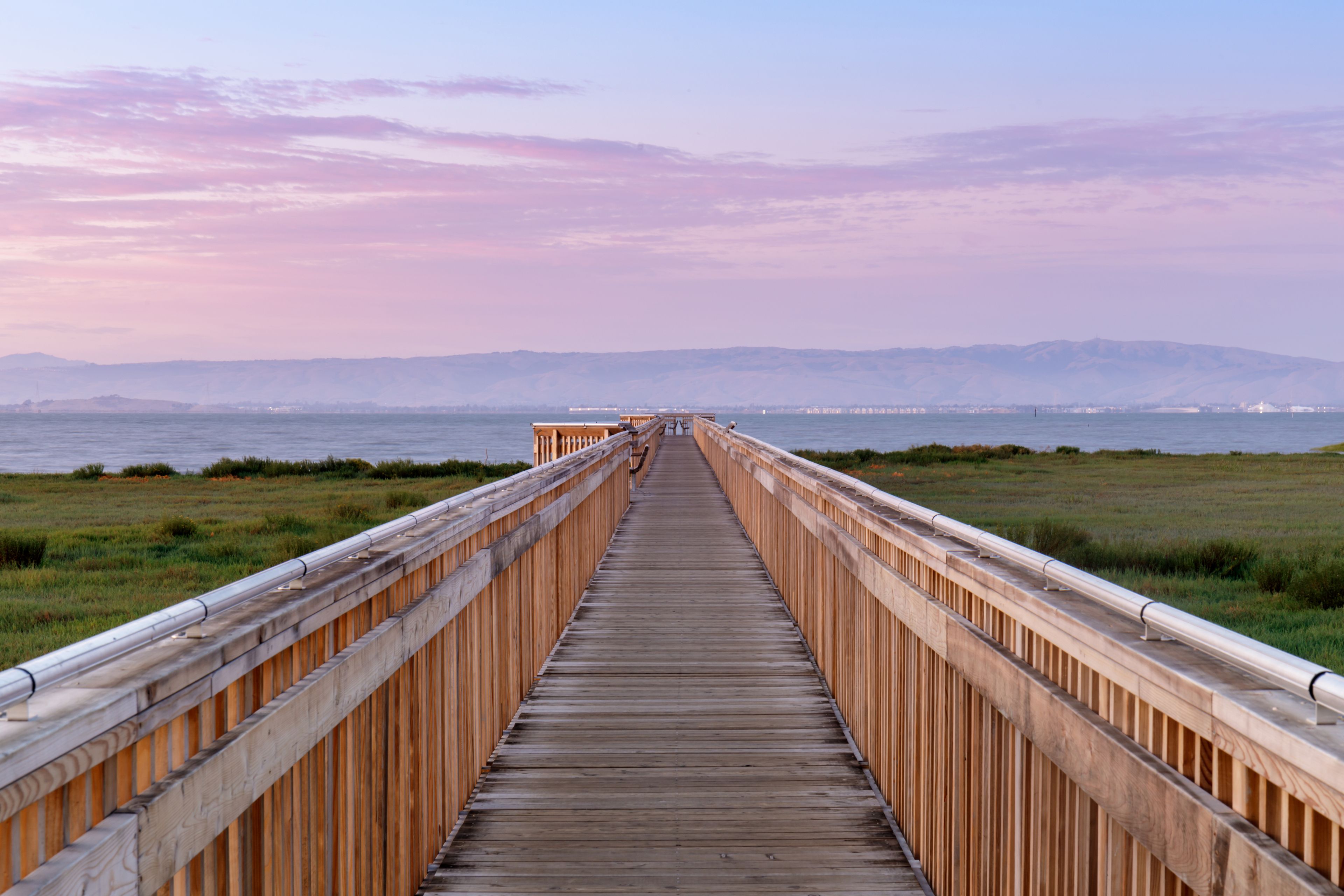 A wooden boardwalk leading to a body of water with distant mountains.