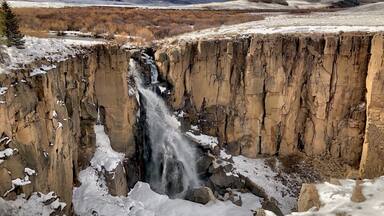 A waterfall cascading down a rocky cliff surrounded by snow-covered terrain and distant mountains.