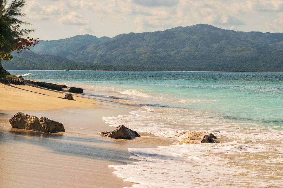 A beach with clear turquoise water, sandy shore, and a mountain range in the background.