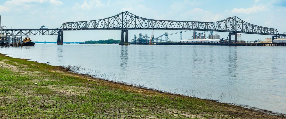 A large steel bridge spanning a river with industrial facilities in the background.