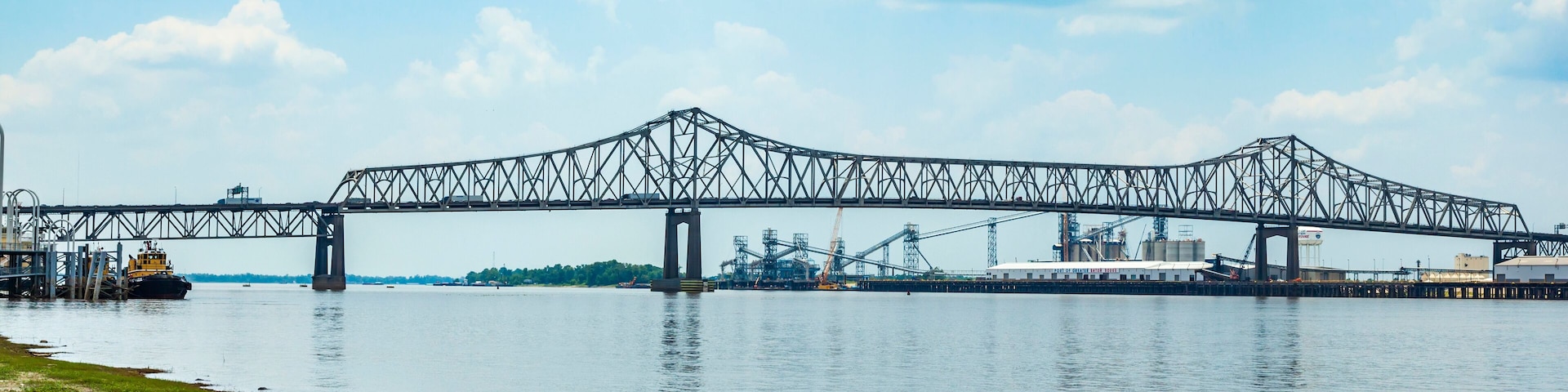 A large steel bridge spanning a river with industrial facilities in the background.