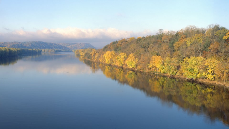 A calm river with autumn foliage reflecting on the water, surrounded by forested hills.