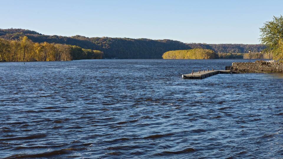 A calm lake with a dock extending into the water, surrounded by autumn-colored trees and hills in the background.