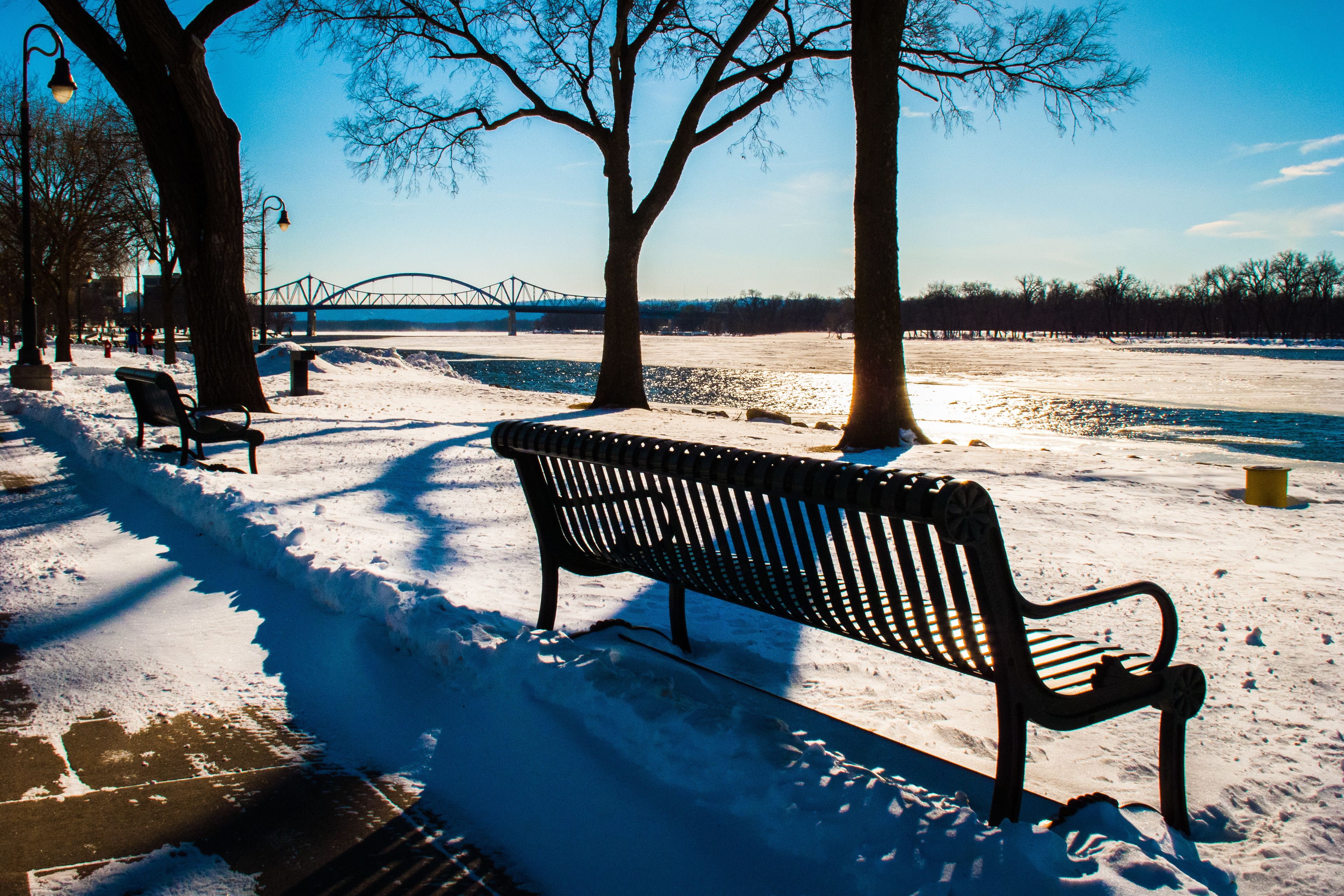 A snow-covered park with benches, trees, a bridge, and a river.