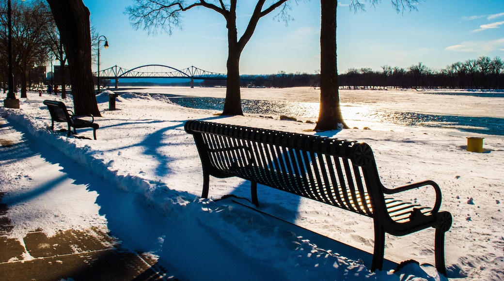 A snow-covered park with benches, trees, a bridge, and a river.
