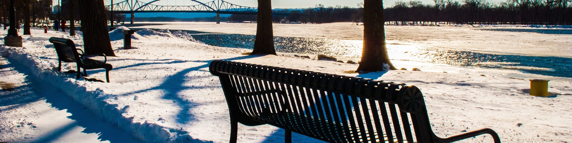 A snow-covered park with benches, trees, a bridge, and a river.
