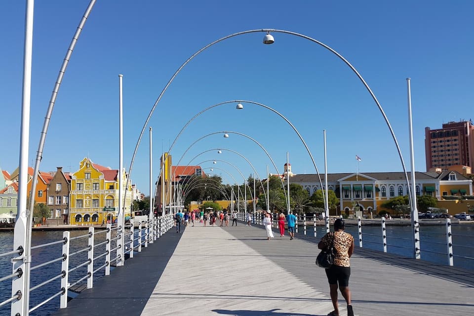 Een voetgangersbrug met gebogen verlichting, kleurrijke gebouwen en een heldere blauwe lucht.