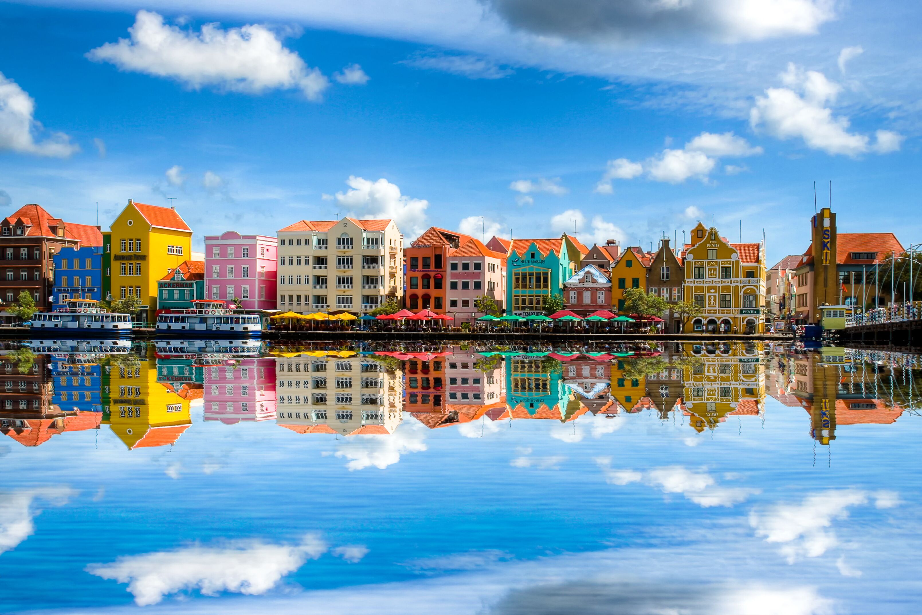 Uma orla com edifícios coloridos refletidos na água, céu azul límpido e mar calmo.