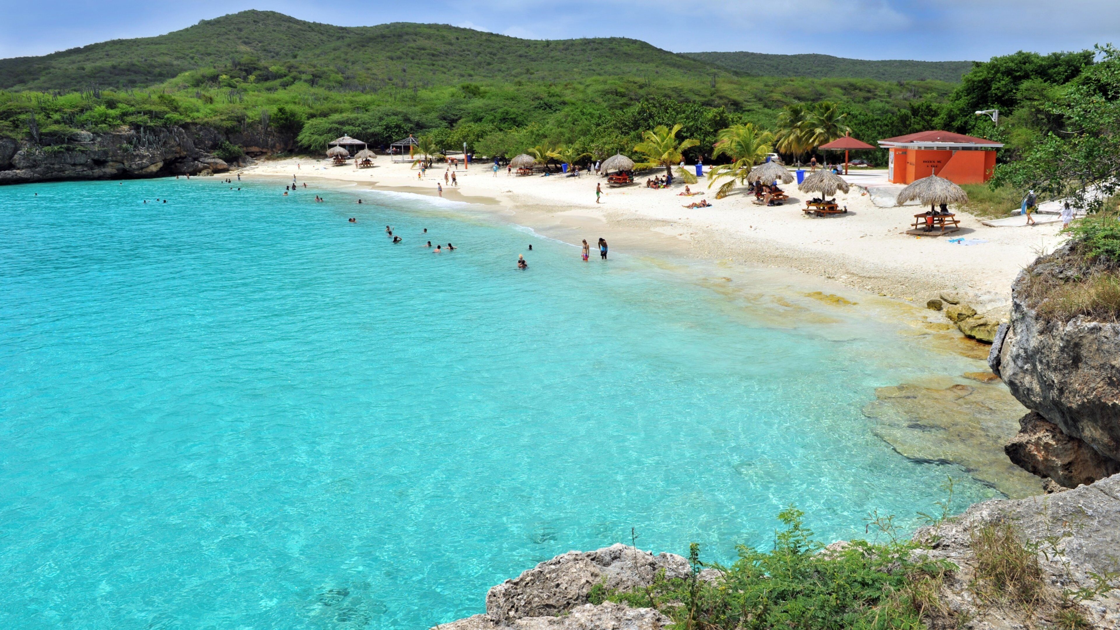 A turquoise beach with clear water, white sand, and lush green hills in the background.
