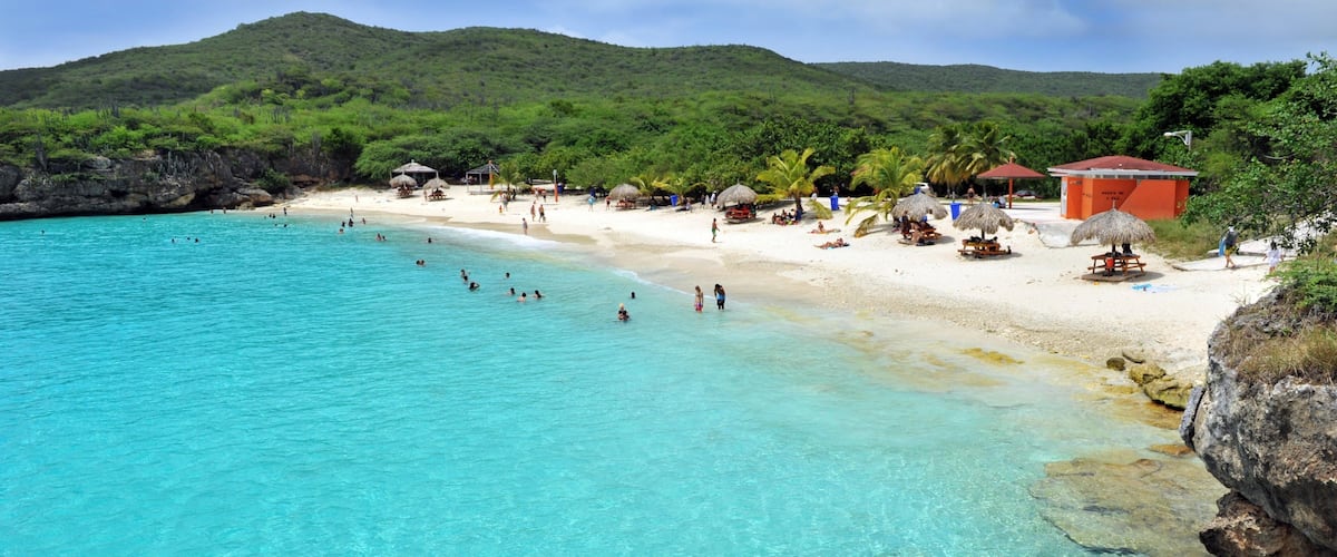 A turquoise beach with clear water, white sand, and lush green hills in the background.