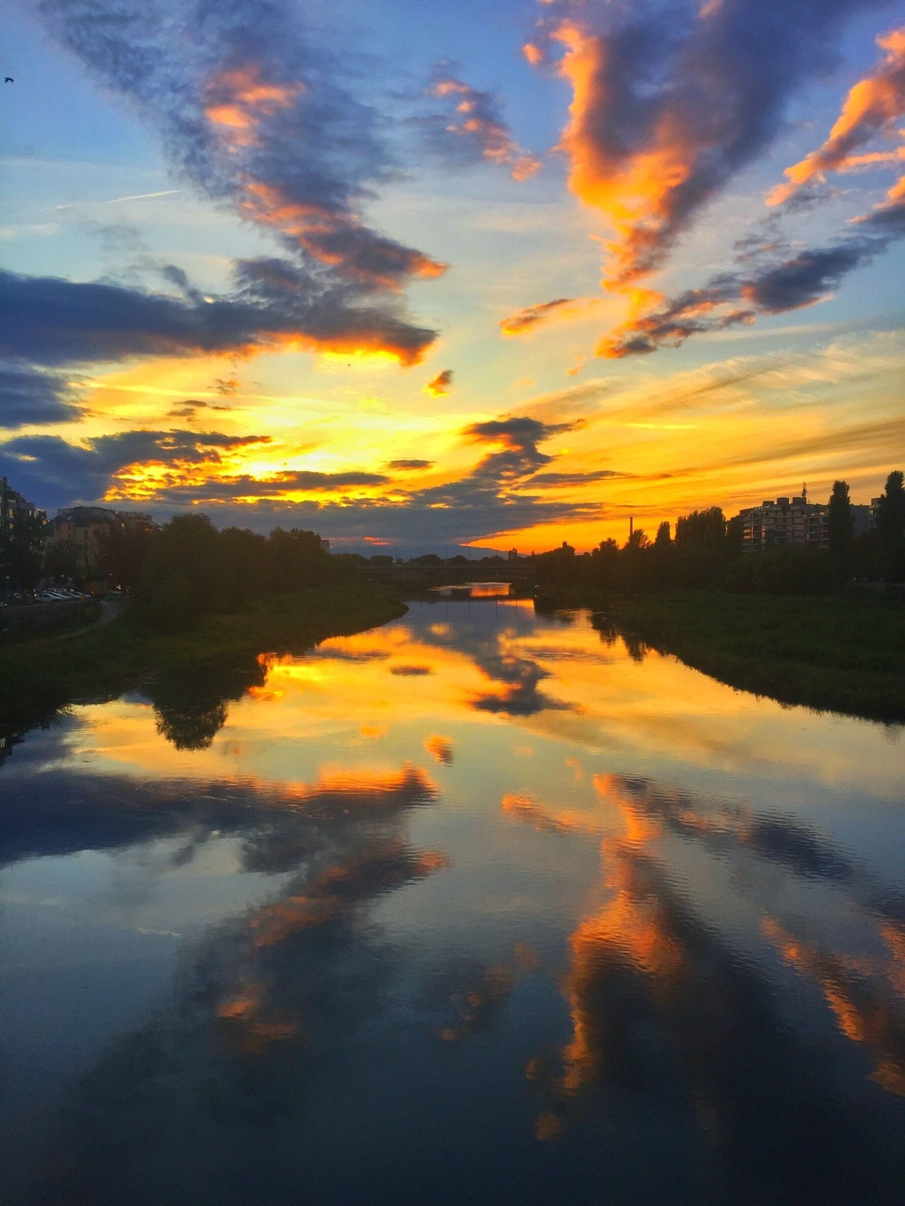 Un atardecer sobre un río tranquilo que refleja nubes coloridas, con edificios y árboles al fondo.
