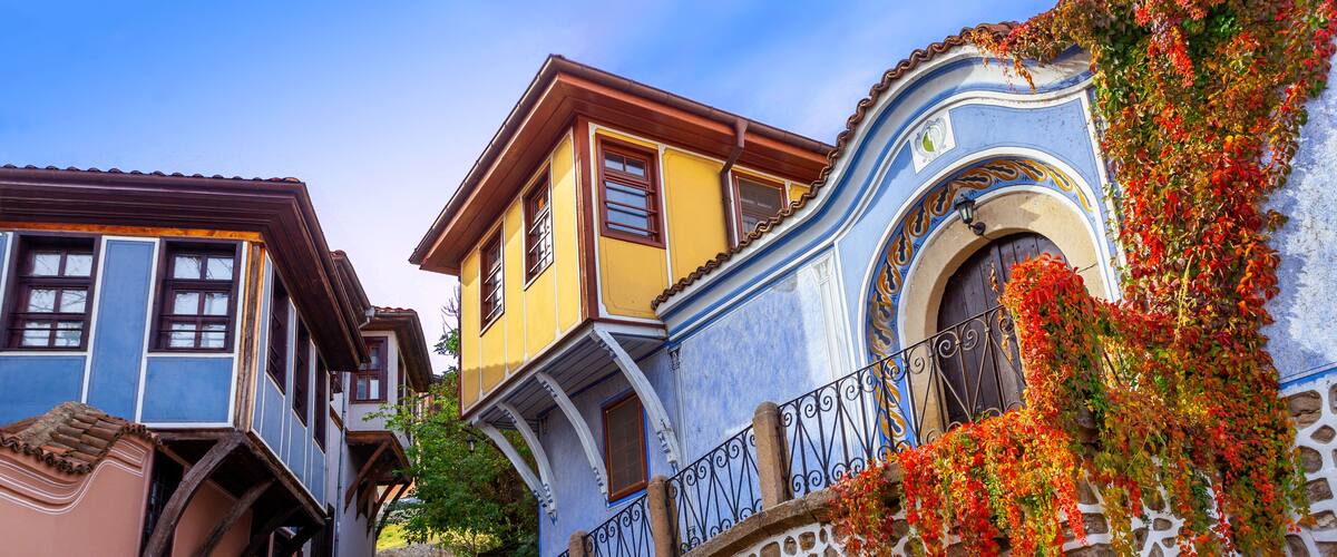A stone staircase leading to colorful buildings with blue and yellow facades, a stone wall with red flowers, and a clear blue sky.