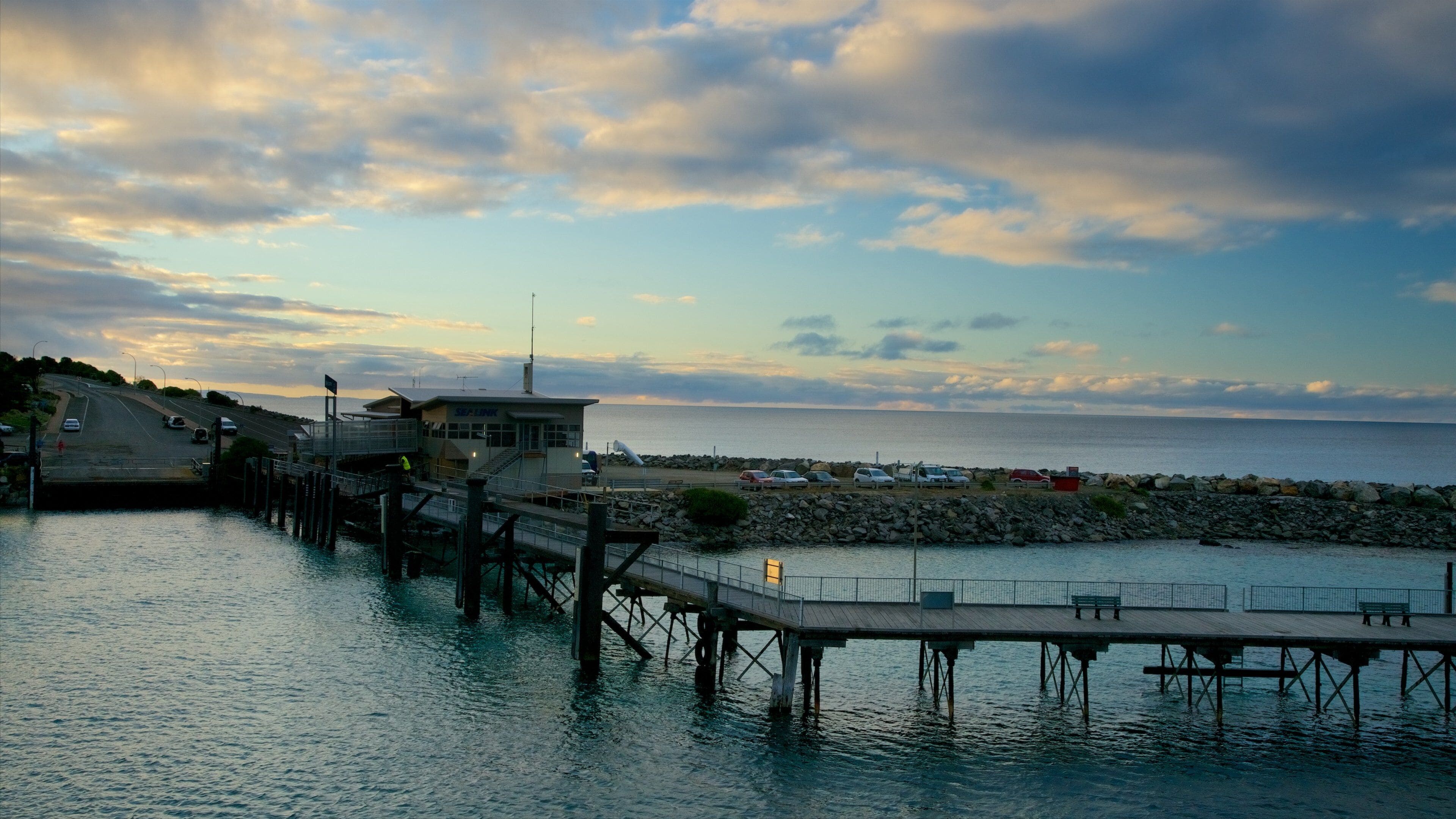 A pier extending into calm waters, a rocky shoreline, and a distant view of the sea under a cloudy sky.