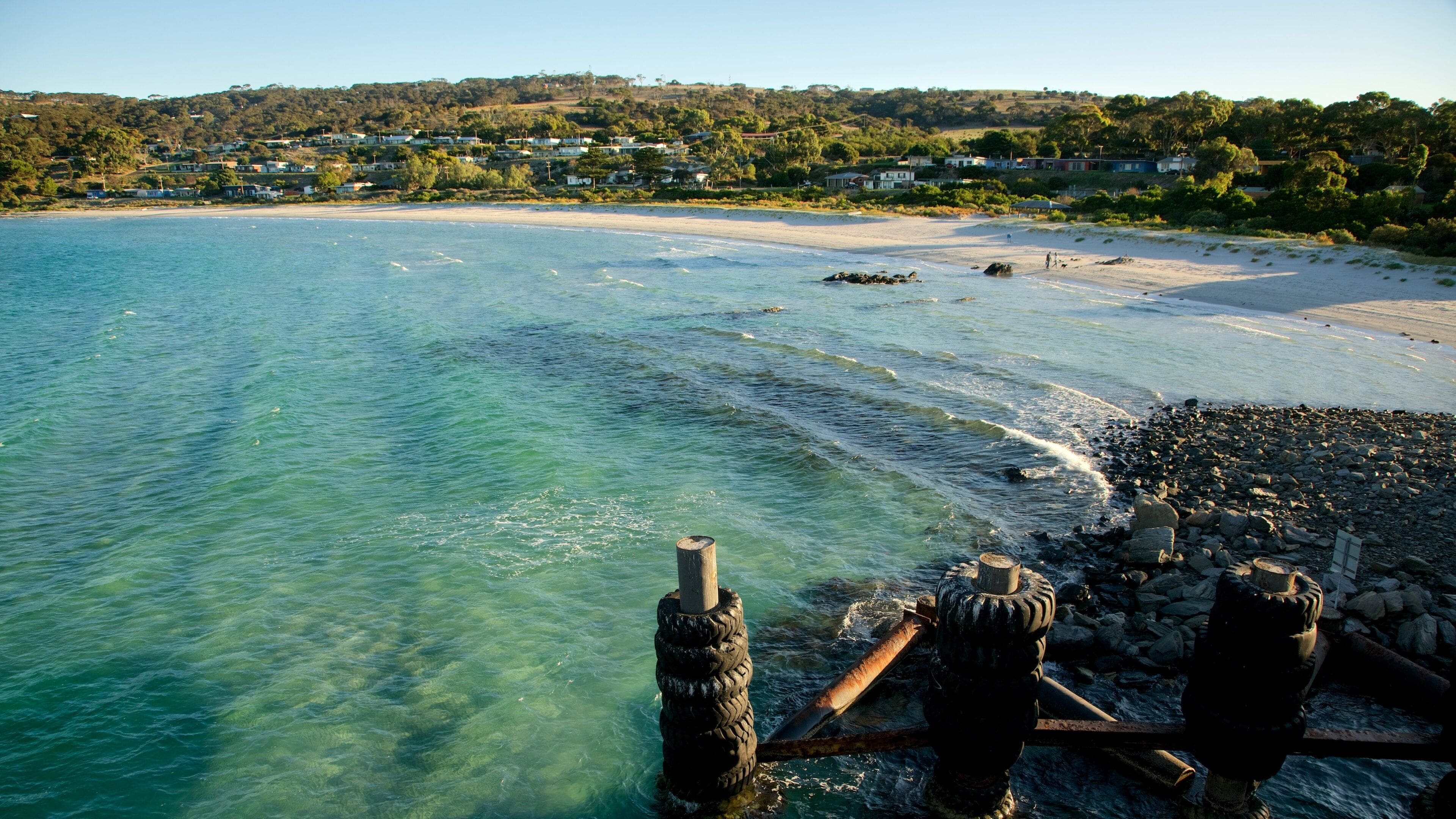 A coastal scene with clear turquoise water, a sandy beach, and a rocky shoreline. In the background, there are houses on a hillside.
