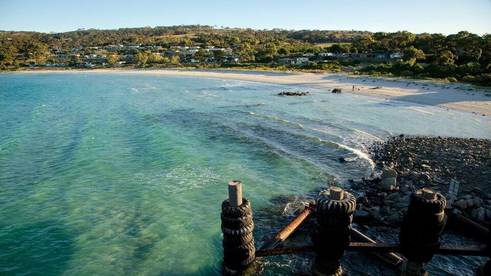 A coastal scene with clear turquoise water, a sandy beach, and a rocky shoreline. In the background, there are houses on a hillside.