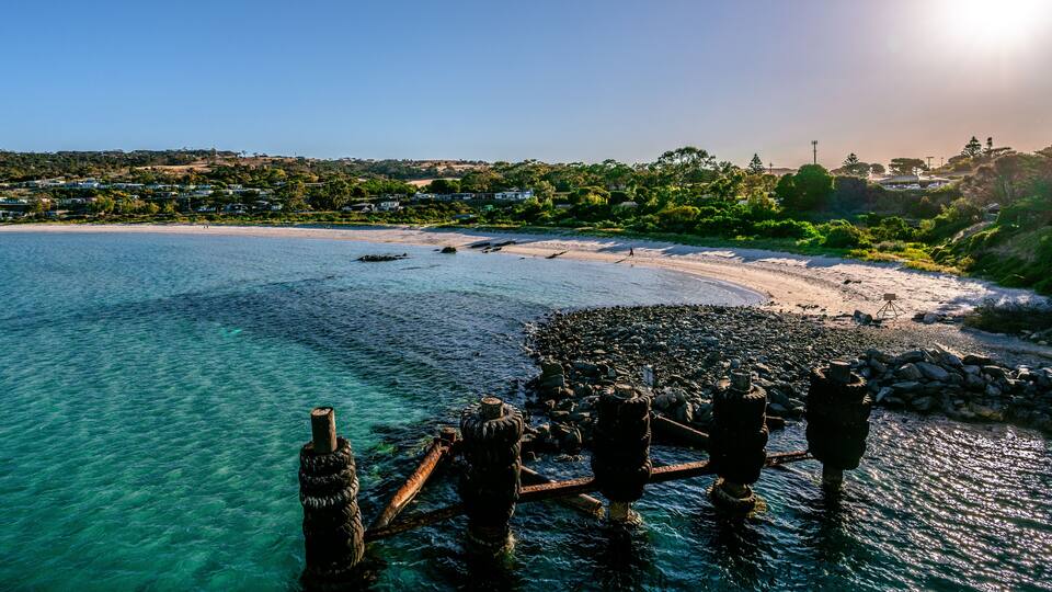 A coastal landscape with clear turquoise waters, a sandy beach, and a rocky outcrop with rusted structures.