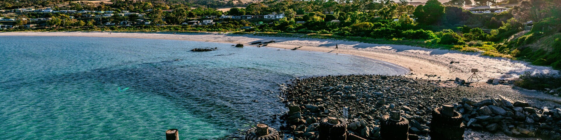 A coastal landscape with clear turquoise waters, a sandy beach, and a rocky outcrop with rusted structures.