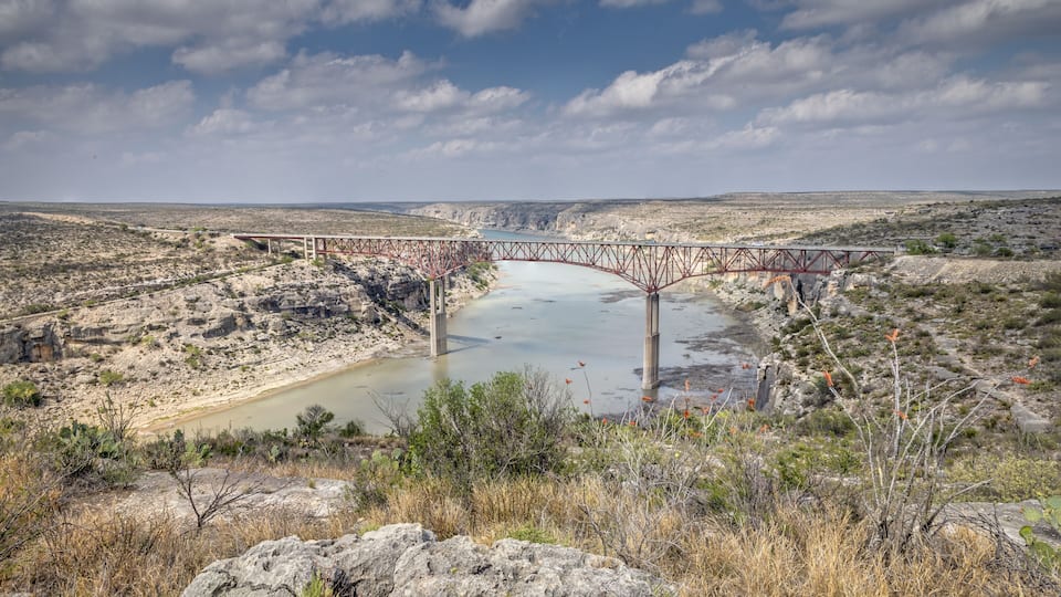 A red metal bridge over a river in a desert landscape.