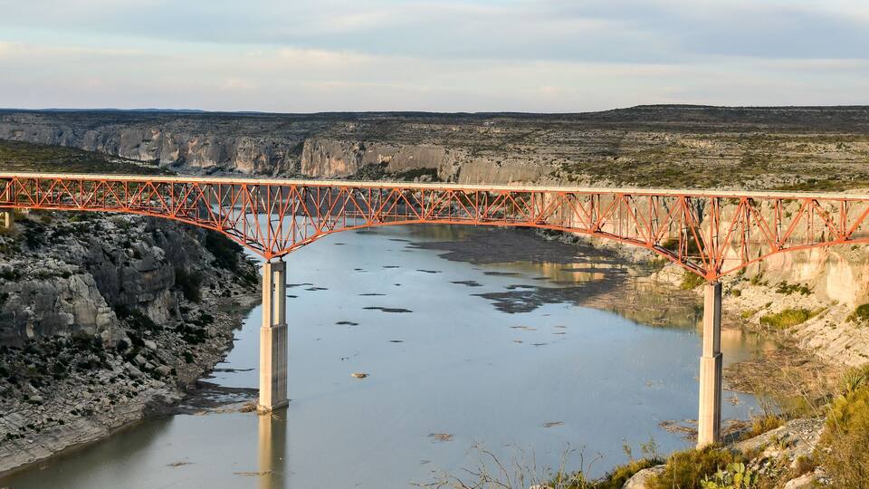 A red bridge over a river with rocky cliffs and sparse vegetation.