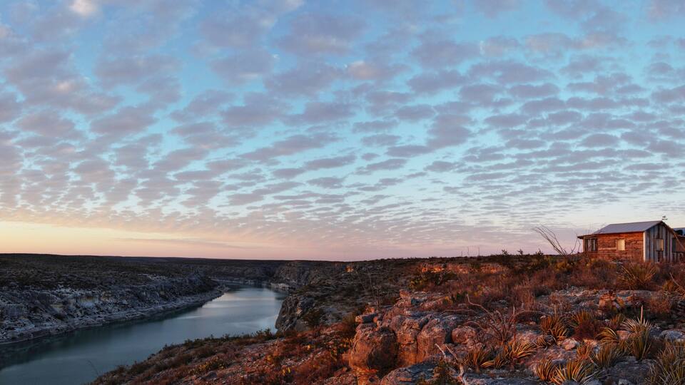 A rugged landscape with a river, rocky terrain, sparse vegetation, and a small wooden structure.