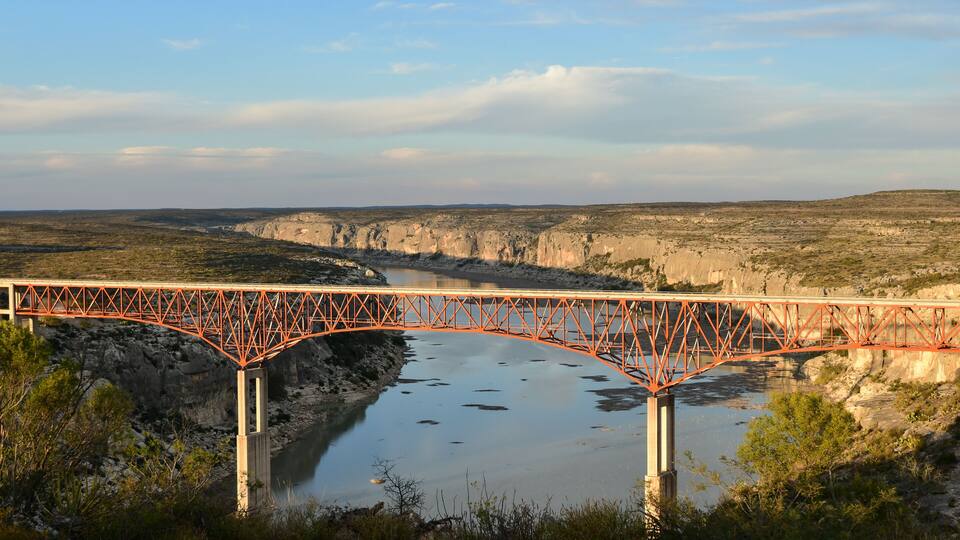 A red metal bridge over a river with cliffs in the background.