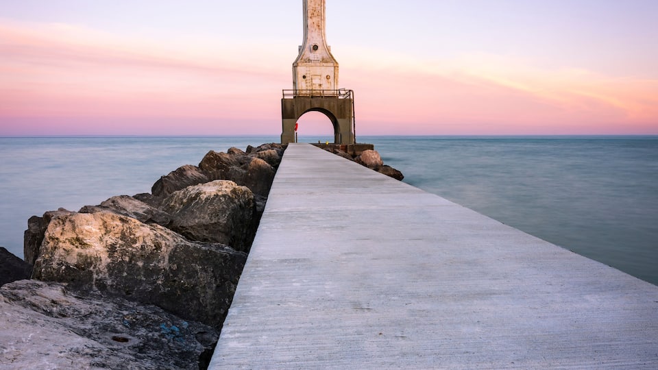 A pier leading to a lighthouse structure with a clear sky and calm sea.