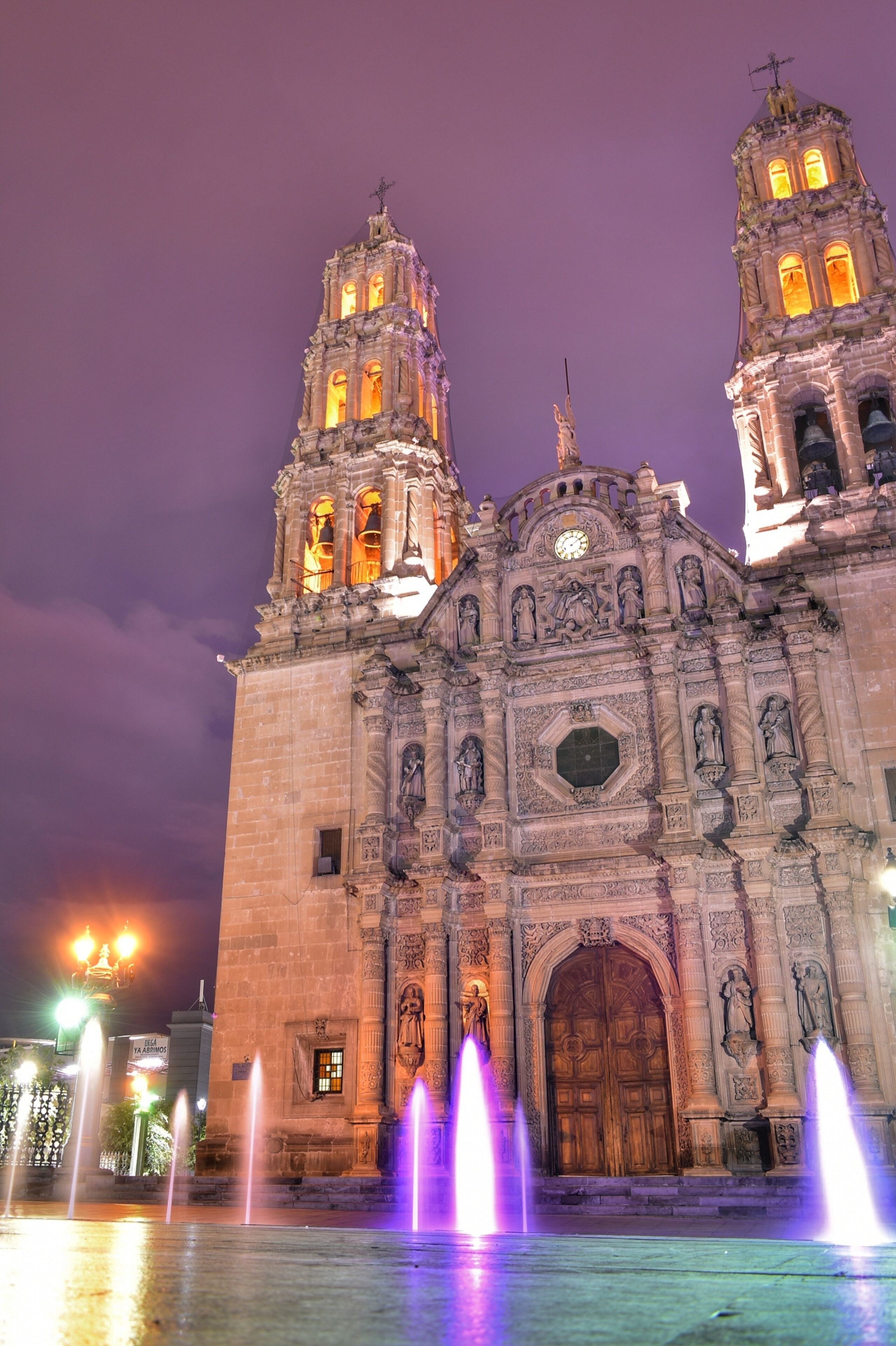 Una catedral histórica con dos torres de campanas, fachada ornamentada y una fuente en primer plano.