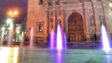 Uma catedral histórica com duas torres sineiras, fachada ricamente decorada e uma fonte em frente.