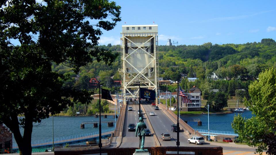 A bridge with a statue in the foreground, a river, and a forested hillside in the background.