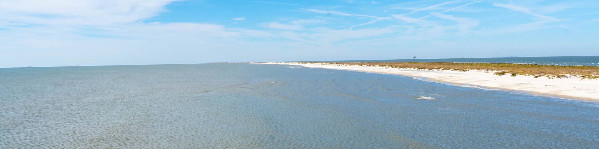 A wide beach with clear water, a sandy shore, and a distant ship.