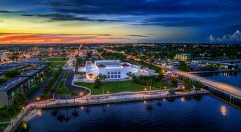 A waterfront cityscape at dusk with a prominent white building surrounded by greenery and a marina.