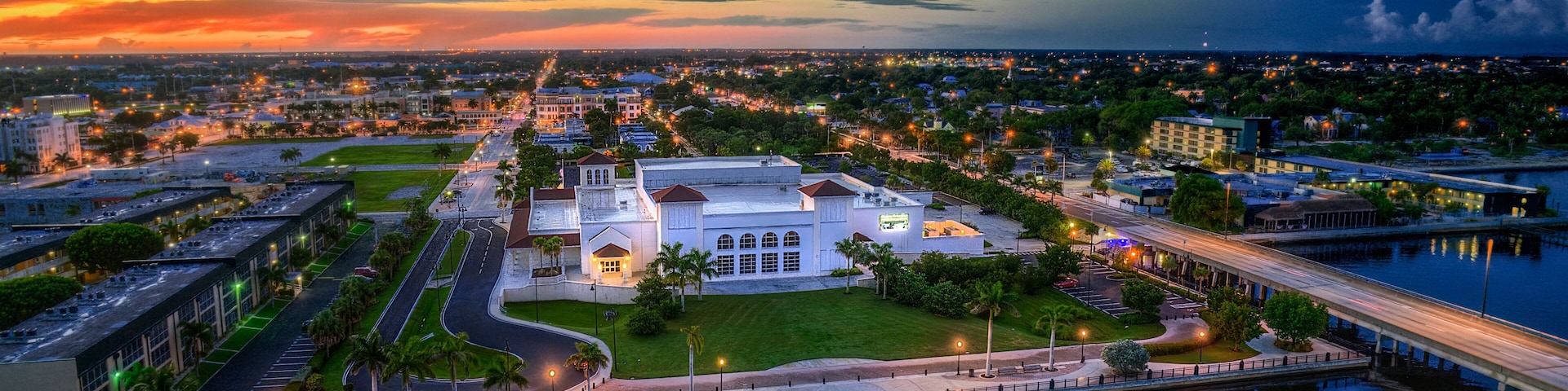 A waterfront cityscape at dusk with a prominent white building surrounded by greenery and a marina.
