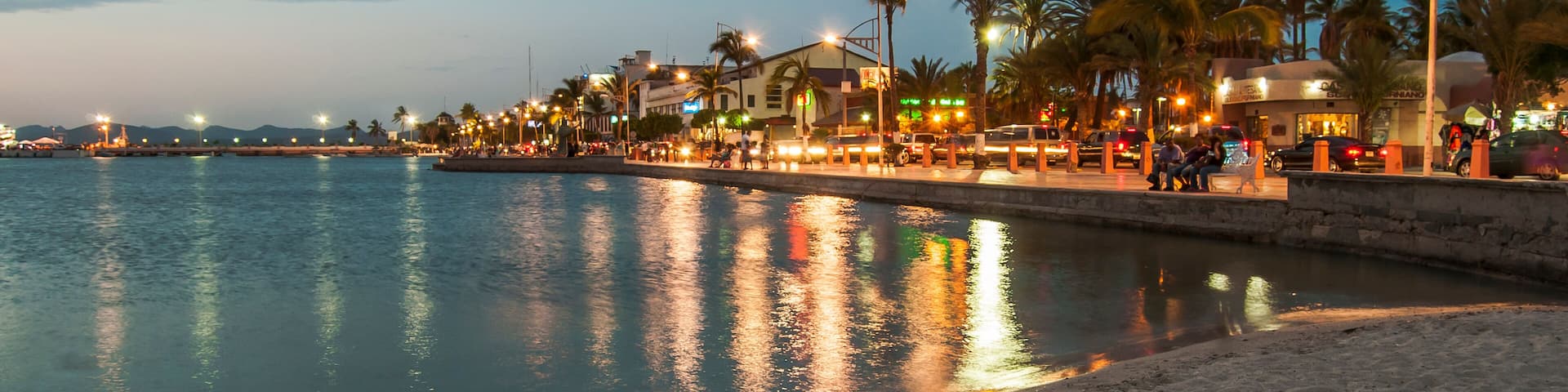 A waterfront promenade with palm trees, street lamps, and buildings reflecting on the water.
