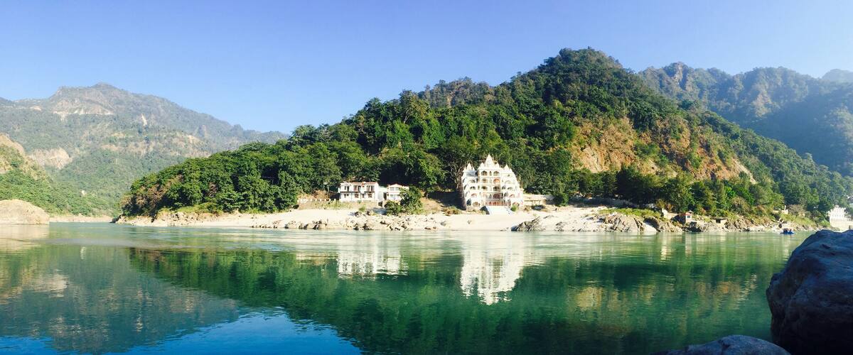 A serene river with clear reflections, a white building with multiple tiers, and a lush green mountain in the background.