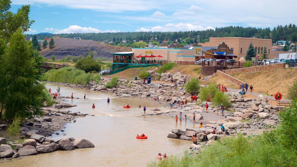 A river with people rafting and walking along the banks. There are buildings and a forested hill in the background.