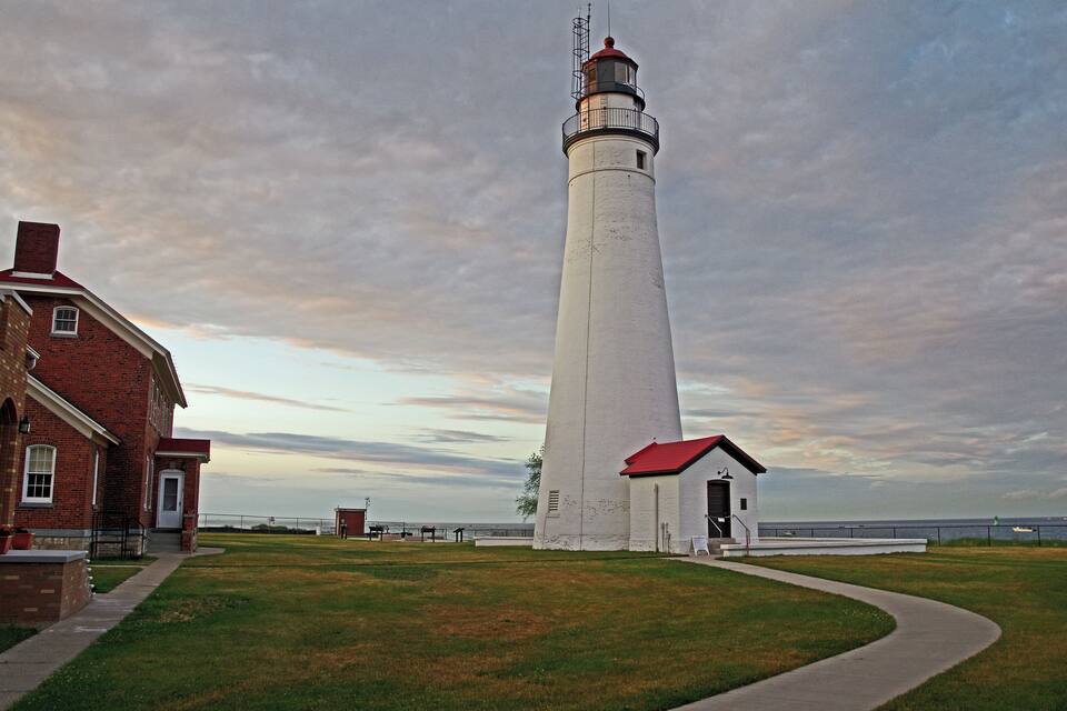 A tall white lighthouse with a red roof, a brick building, and a grassy area with a path leading to the lighthouse.