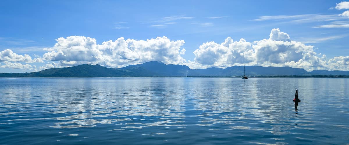 A calm lake with a sailboat, mountains in the background, and a clear sky with clouds.