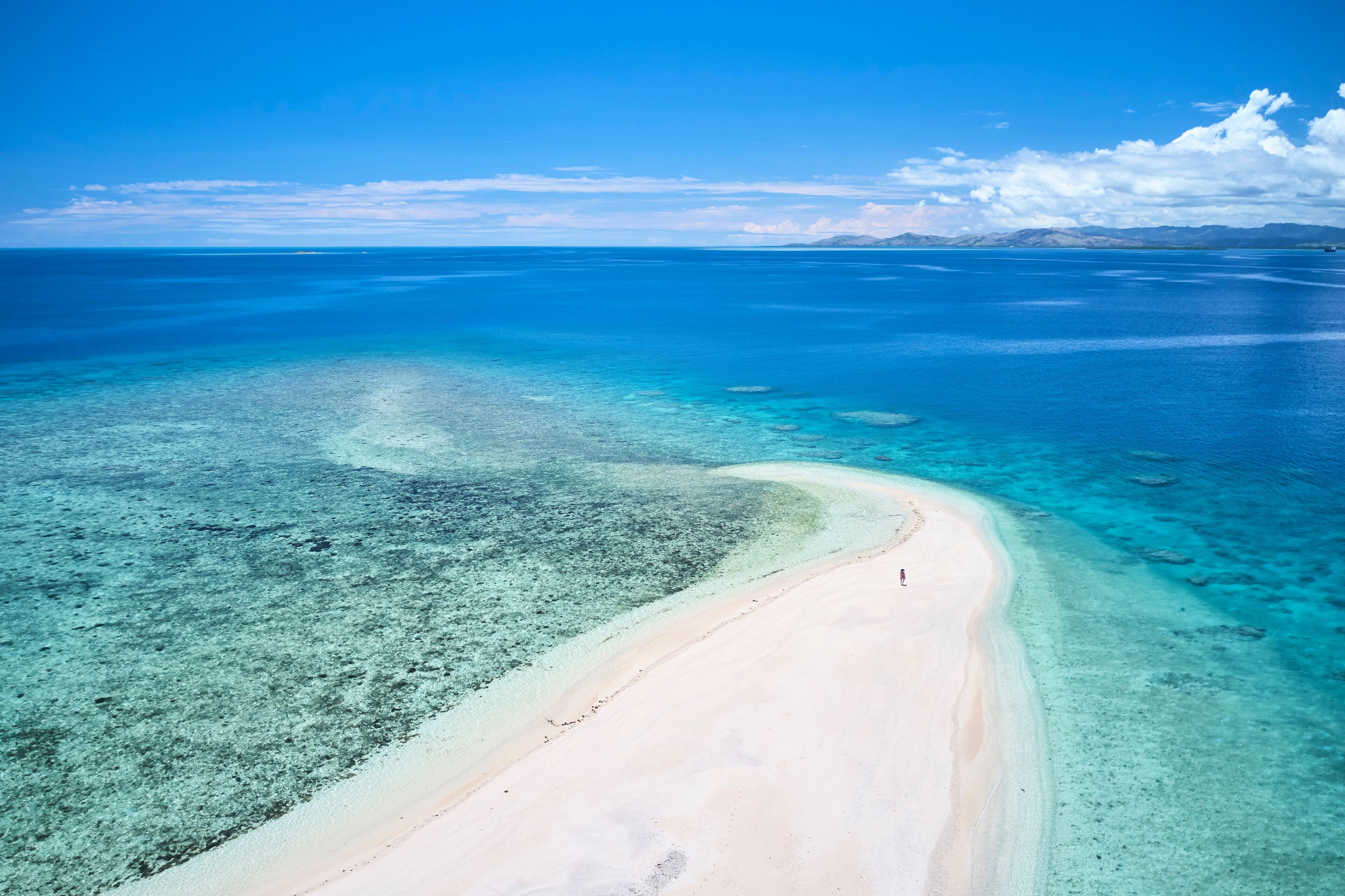 Mar azul límpido, areal arenoso e montanhas ao longe.