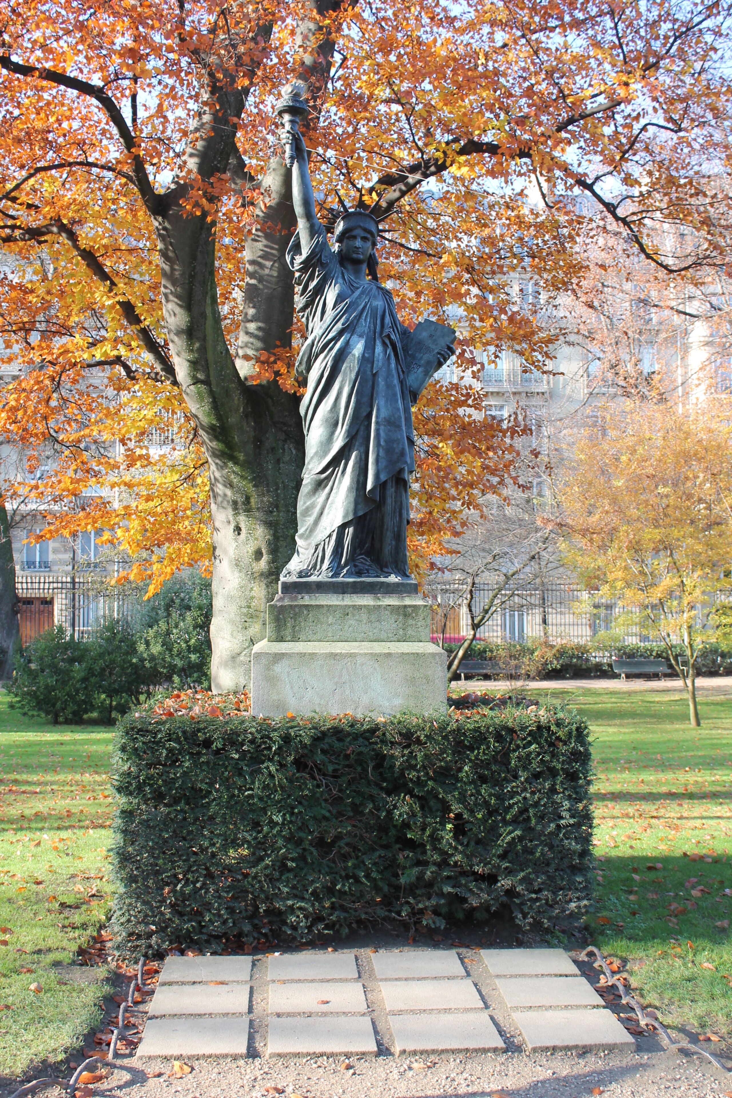 A statue of a figure holding a torch, surrounded by autumn leaves and a hedge.