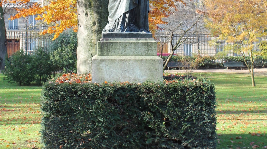 A statue of a figure holding a torch, surrounded by autumn leaves and a hedge.