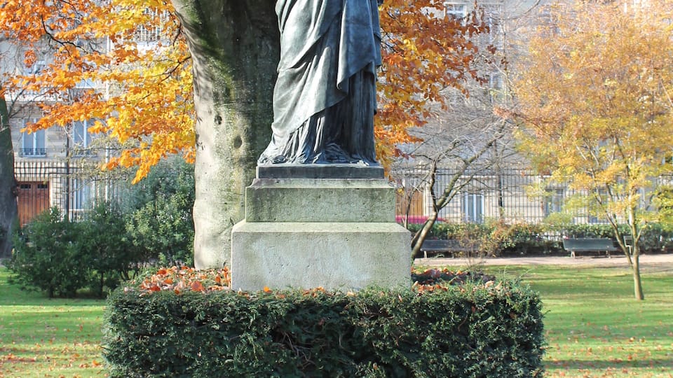 A statue of a figure holding a torch, surrounded by autumn leaves and a hedge.
