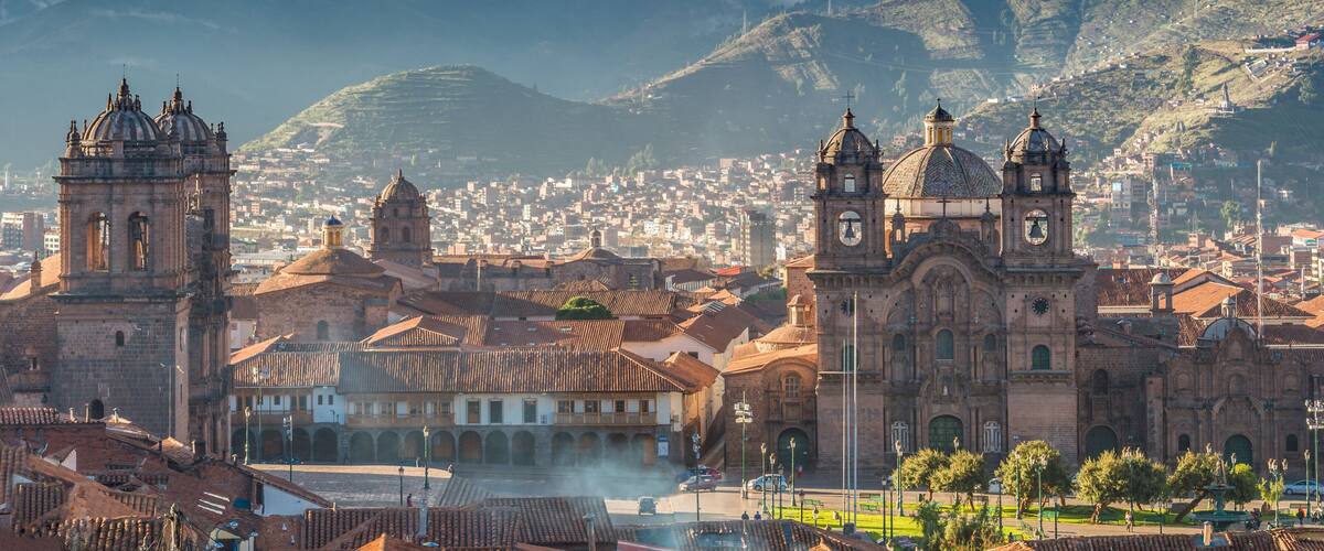A historic city with prominent cathedral towers, a bustling square, and a backdrop of mountains and clouds.