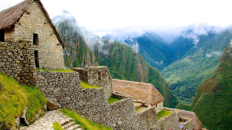 Ancient stone ruins with thatched roofs, set against a backdrop of lush green mountains and misty peaks.