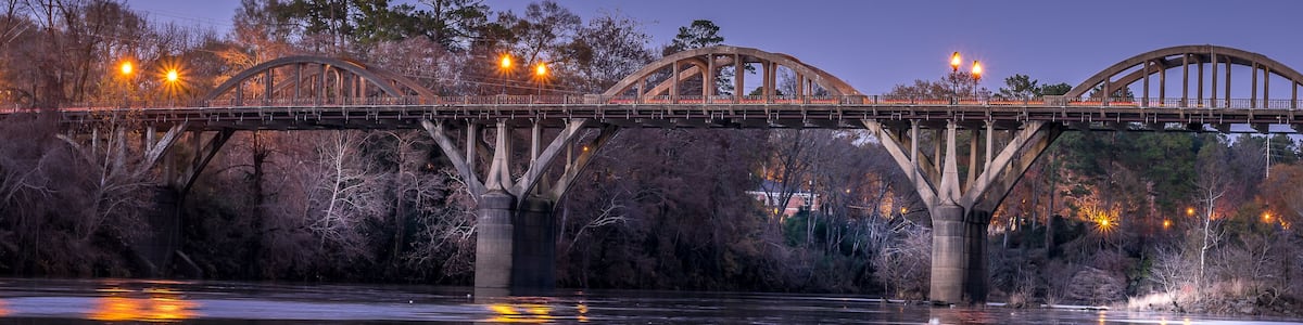 A bridge over a calm river at dusk with lights reflecting on the water.