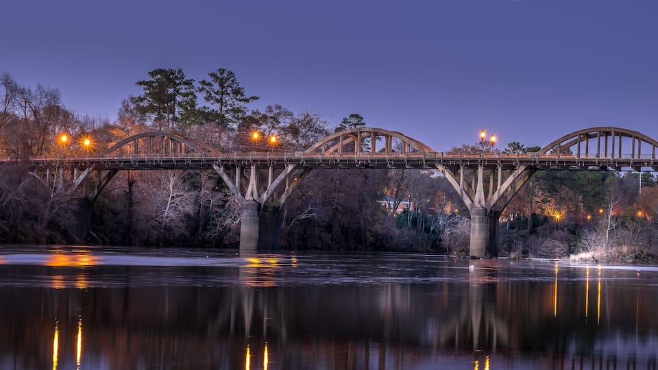 A bridge over a calm river at dusk with lights reflecting on the water.