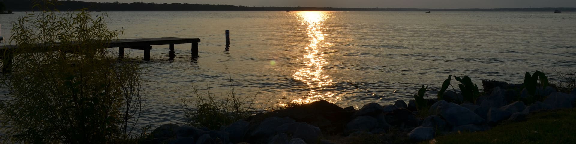 A sunset over a calm lake with a wooden dock extending into the water, surrounded by rocks and vegetation.