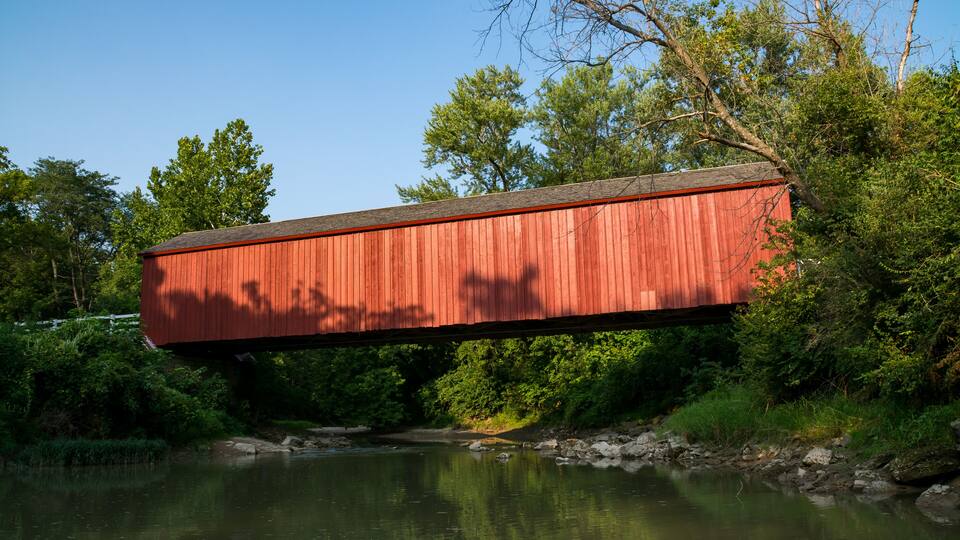 A red covered bridge over a calm river, surrounded by lush greenery and trees.