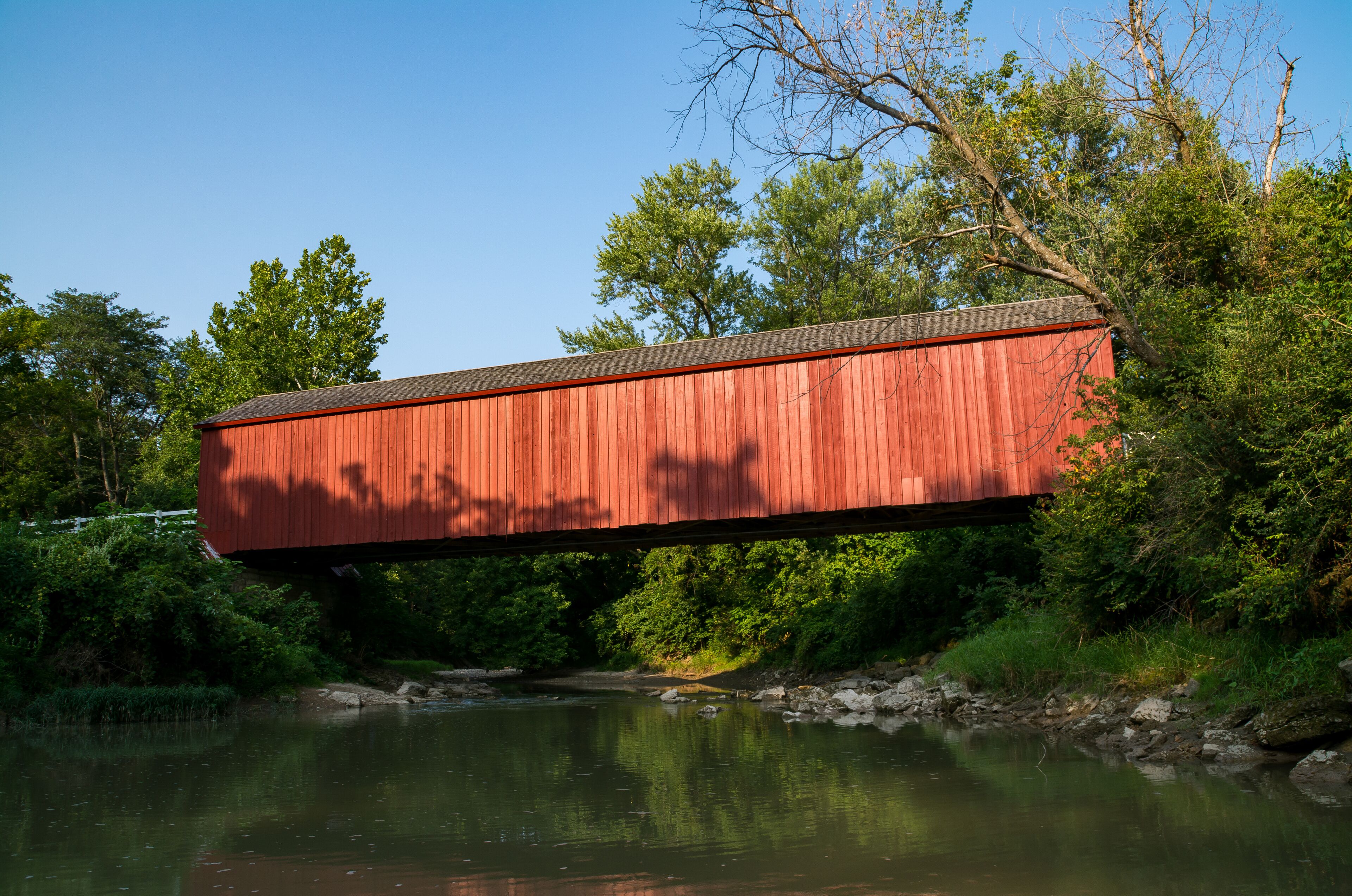 A red covered bridge over a calm river, surrounded by lush greenery and trees.