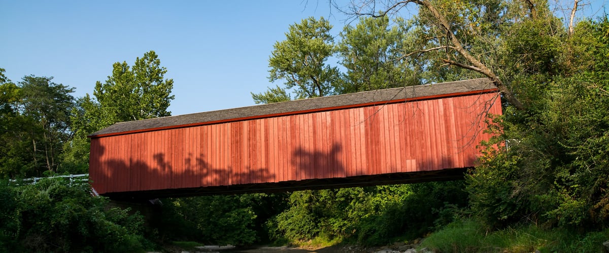 A red covered bridge over a calm river, surrounded by lush greenery and trees.