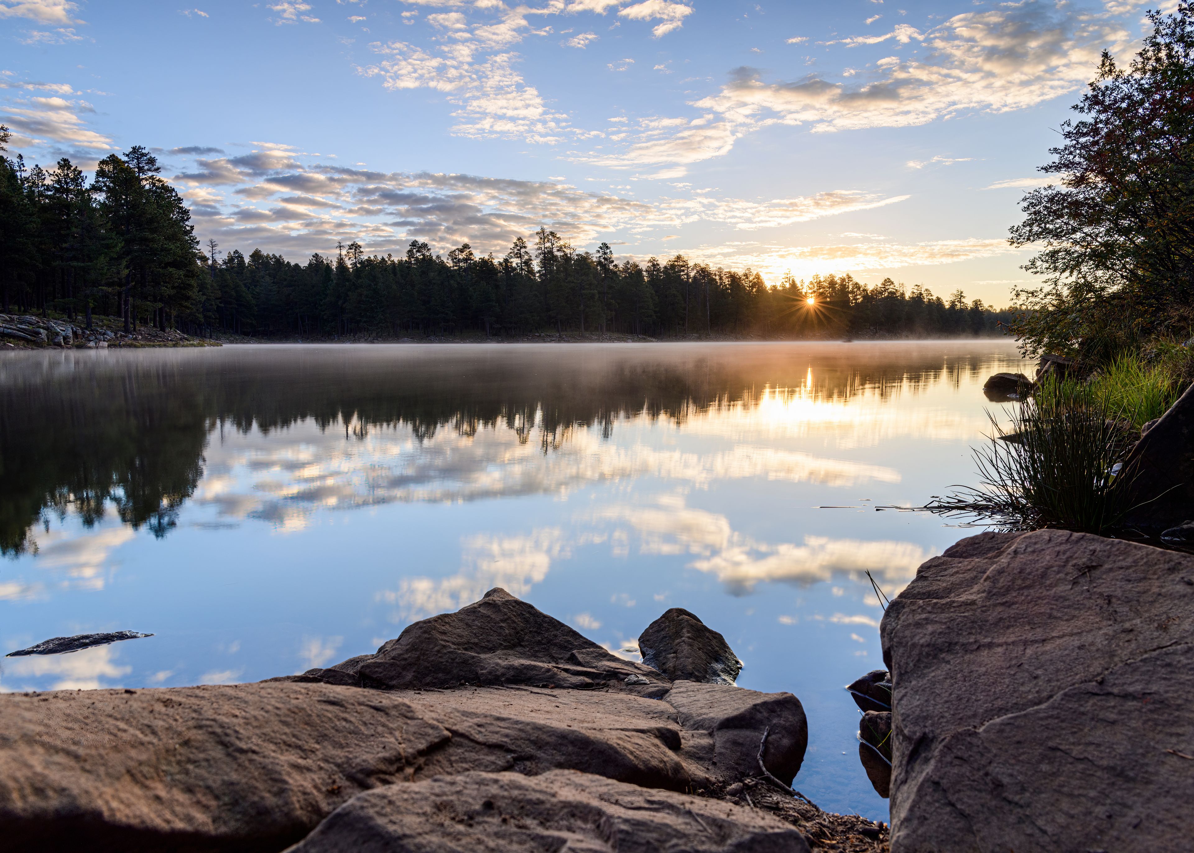 A calm lake with a reflection of trees and sky, surrounded by rocks and a forest.